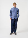 A male model poses against a light grey studio backdrop wearing the Indigo Prince Chambray Daybrook Shirt. The classic long-sleeve button-down shirt features a point collar and front patch pocket, worn unbuttoned over a plain white tee.