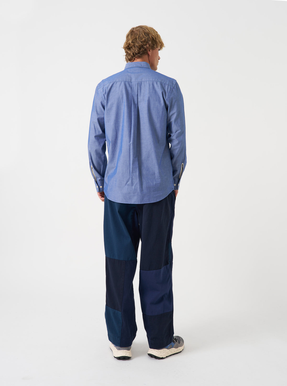 Back view of a male model wearing the Indigo Prince Chambray Daybrook Shirt against a plain light grey studio backdrop. The back panel and long sleeves of the indigo blue chambray shirt are prominently shown.
