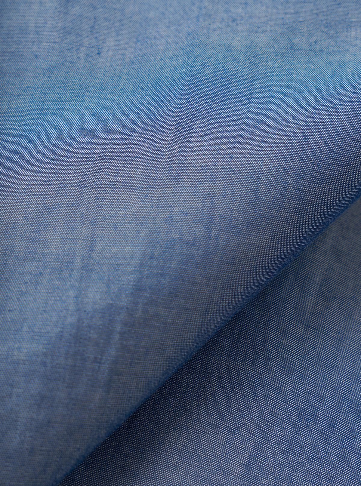 Close-up of the indigo chambray fabric used in the Daybrook shirt, showing a fine two-tone weave of blue and white threads. A diagonal fold runs across the frame, highlighting the fabric's soft drape and subtle texture variation across the surface.