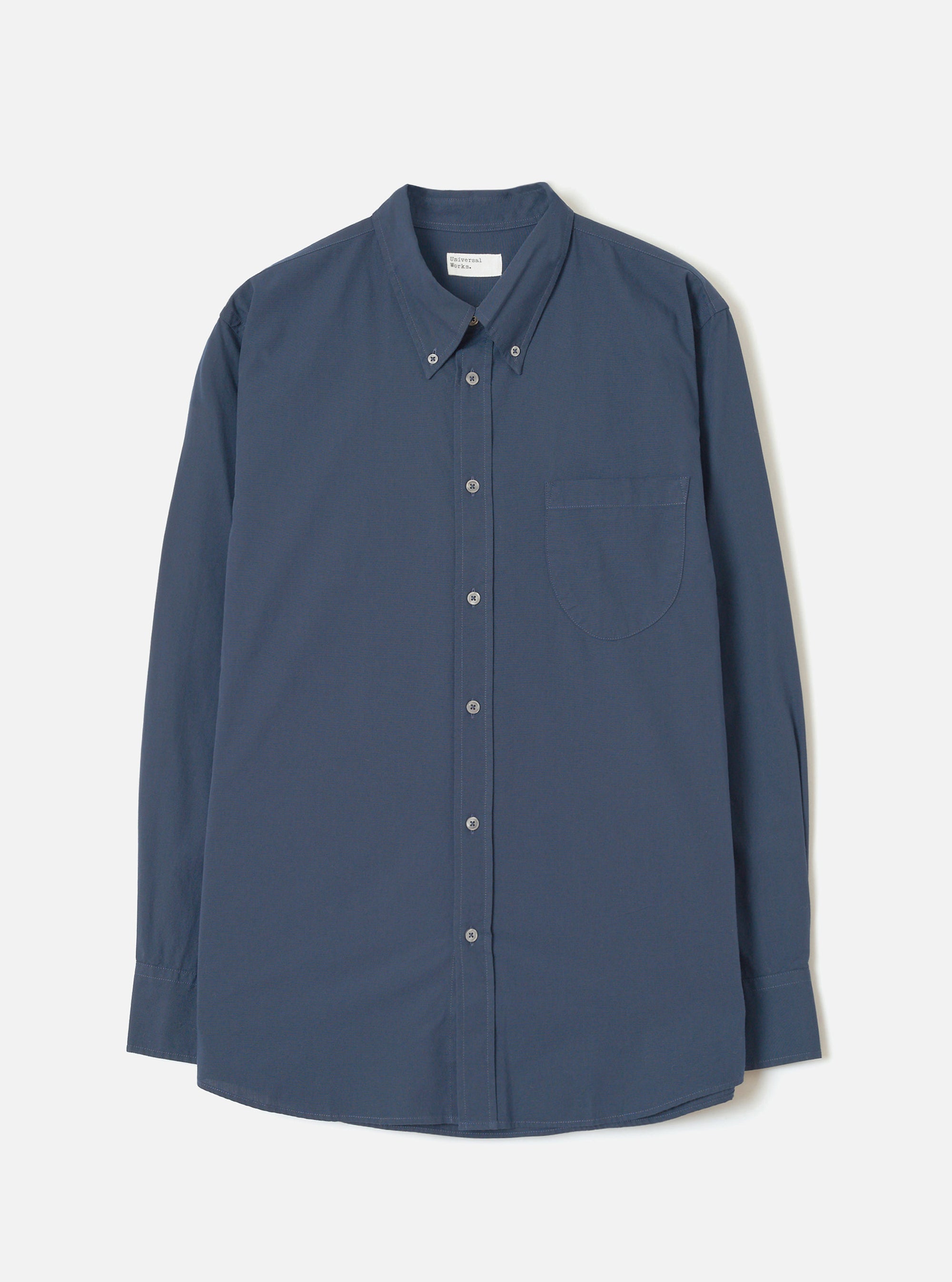 Navy chambray long-sleeve shirt laid flat on a light grey studio backdrop. Features a button-down collar, buttoned placket with six grey buttons, a curved rounded hem, single chest patch pocket, and single-button barrel cuffs. A branded woven label sits at the centre back neck.