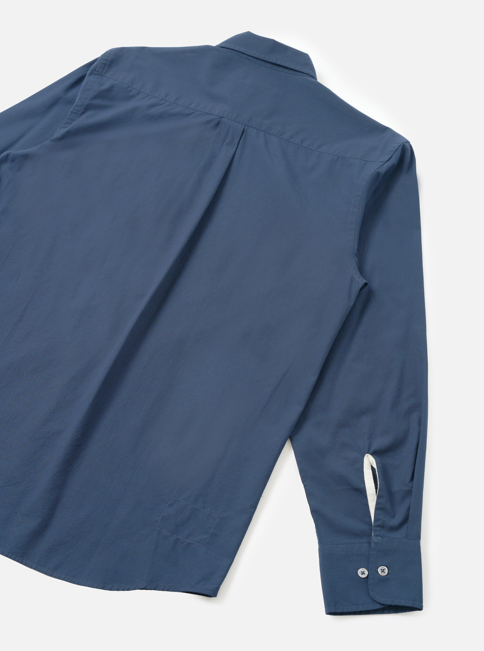 Navy chambray Daybrook shirt laid flat on a white surface, shot from above at an angle showing the back. A box pleat runs from the collar yoke. The long sleeve cuff features two buttons and a cream contrast lining is visible at the opening. Smooth, lightweight fabric in a plain navy weave.