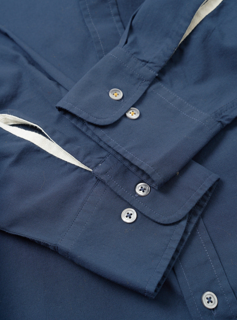 Close-up flat lay of the navy chambray Daybrook shirt showing two buttoned barrel cuffs with silver four-hole buttons. A cream cotton twill tape trim is visible along the placket edge. The plain-weave fabric has a fine texture. Top-stitching details the cuff edges. Shot on a white studio surface.