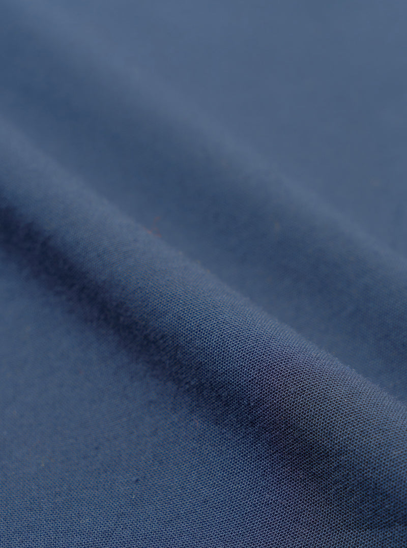 Close-up of navy blue chambray fabric, showing a plain weave texture with a subtle two-tone woven finish. The cloth is folded to create a diagonal ridge across the frame, with soft shadows highlighting the fabric weight and drape. No garment details or background are visible.