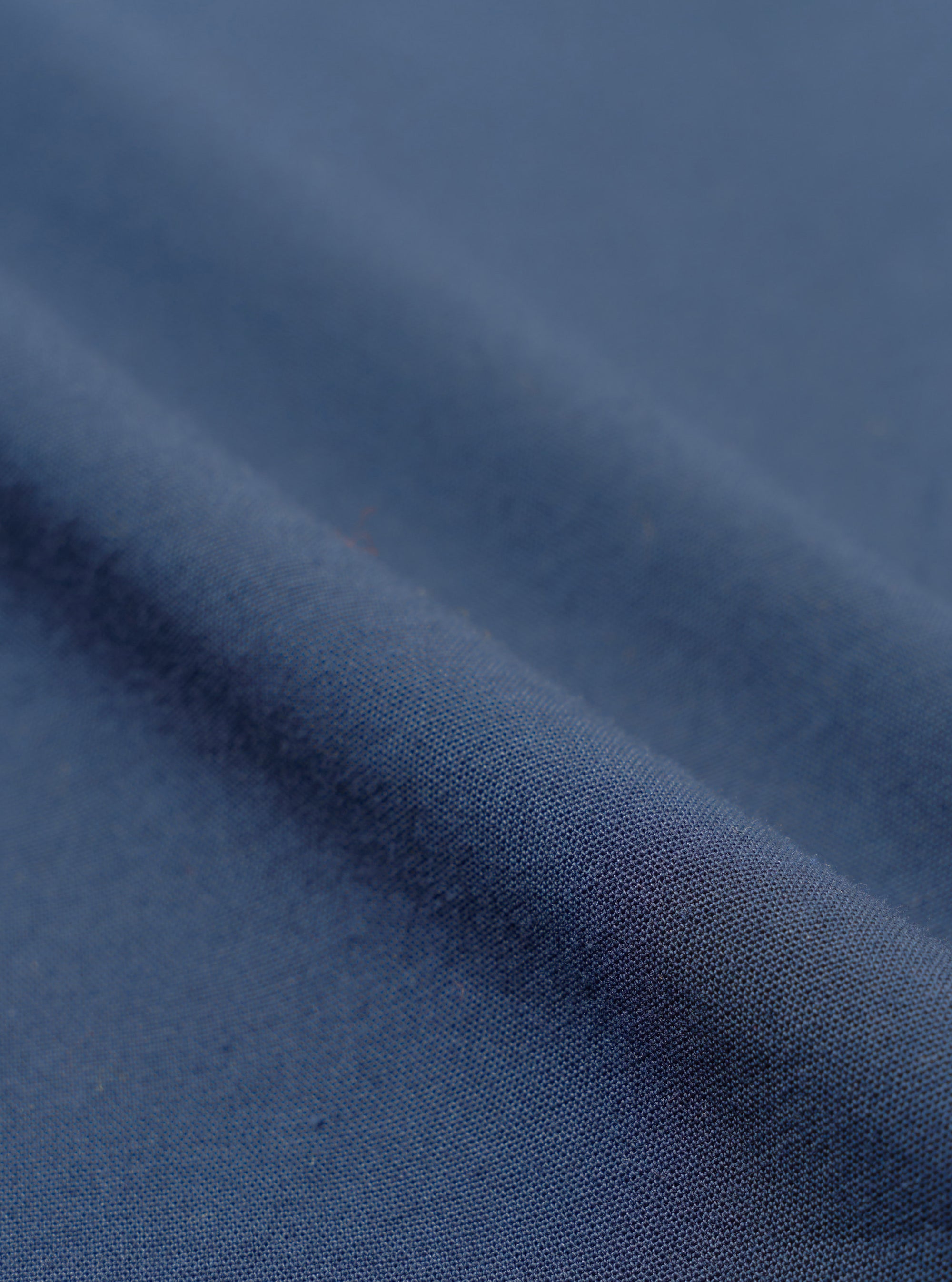 Close-up of navy blue chambray fabric, showing a plain weave texture with a subtle two-tone woven finish. The cloth is folded to create a diagonal ridge across the frame, with soft shadows highlighting the fabric weight and drape. No garment details or background are visible.