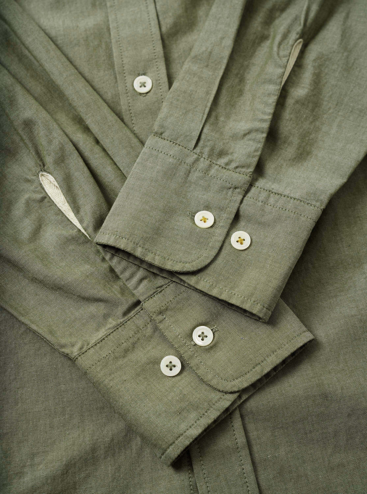 Close-up flat-lay detail of the olive chambray Daybrook shirt, showing two barrel cuffs each fastened with two white four-hole buttons with gold stitching. The placket is visible at the top with a single button. Fine topstitching runs along the cuff edges. The woven chambray fabric has a subtle two-tone texture.