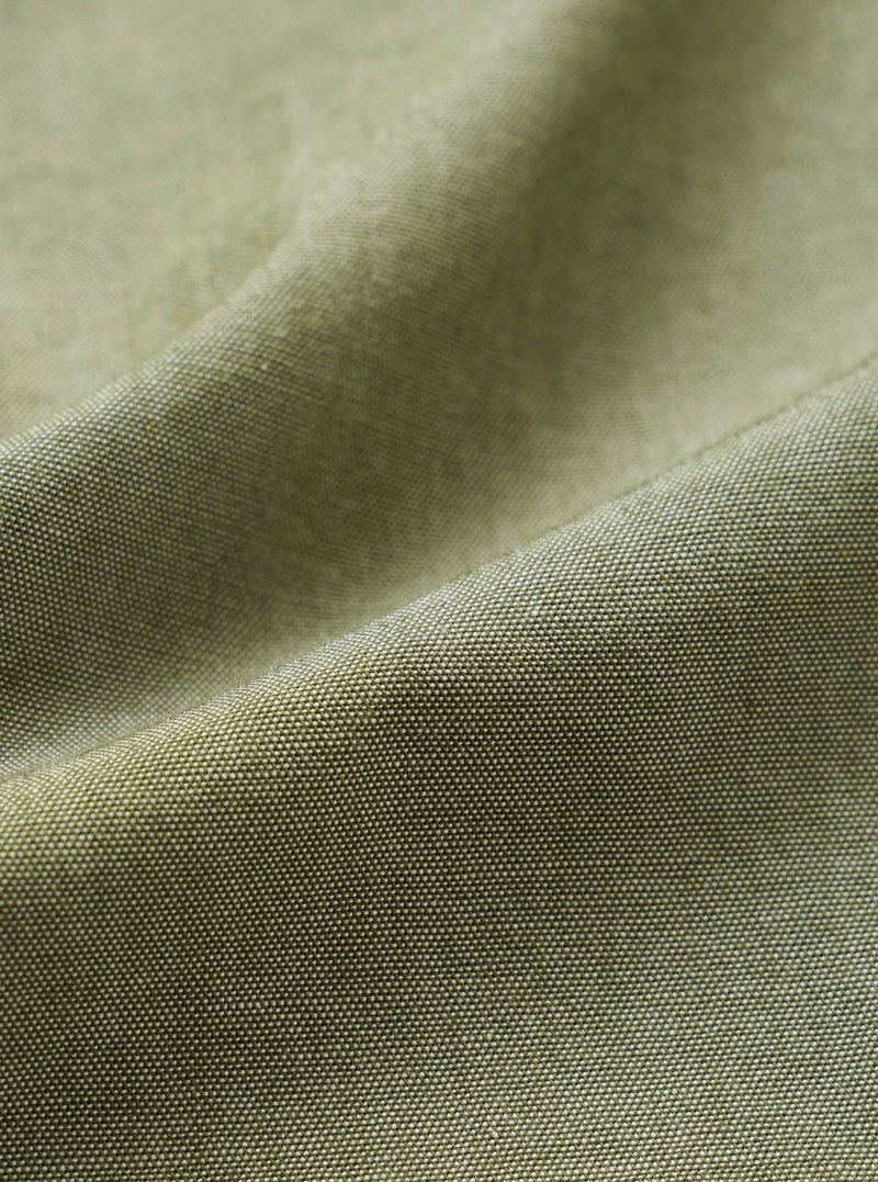 Close-up of the olive green chambray fabric of the Daybrook Shirt, showing a plain weave texture with a subtle two-tone yarn construction. A folded seam runs diagonally across the frame, highlighting the fabric weight and finish.
