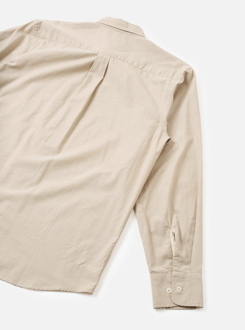 Flat lay of a sand-coloured chambray Daybrook shirt shot from above on a white surface, showing the back and one sleeve. The back features a centre pleat and a small locker loop at the collar. The long sleeve ends in a buttoned cuff with two cream buttons and a placket vent. The spread collar is visible at the top. Fabric has a fine, slightly textured weave.