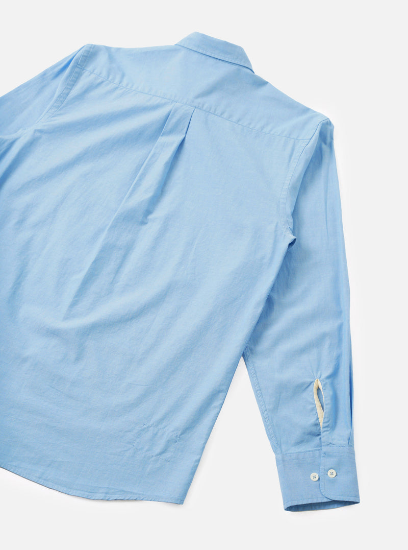 Back view of the sky blue chambray Daybrook shirt laid flat on a white studio backdrop. The plain-weave cotton fabric has a fine, slightly textured finish. Visible details include a back yoke with a central pleat, a spread collar, one long sleeve with a button cuff fastened with two white buttons, and a cream loop tab at the cuff opening.