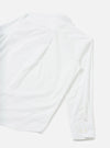 White chambray shirt laid flat on a light grey seamless surface, shot from above at an angle. The image shows the back of the shirt with one long sleeve extending to the lower right. A small collar is visible at the top. The buttoned cuff features two small white buttons. The fabric has a smooth, fine weave texture.