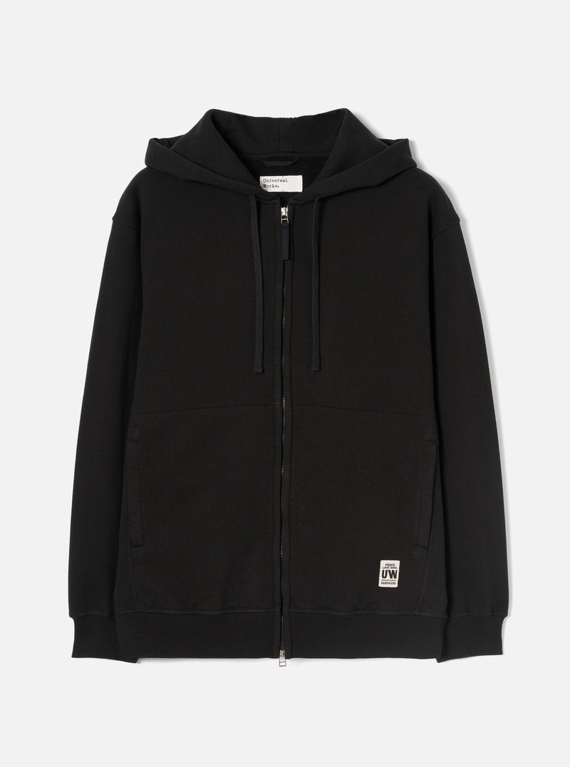 Black zip-through hoodie in brushback sweat fabric, laid flat on a white studio backdrop. Features a jersey-lined hood with navy drawcords, a full-length metal zip, two large patch pockets, and ribbed cuffs and hem. A small woven UW brand label sits at the lower right front.