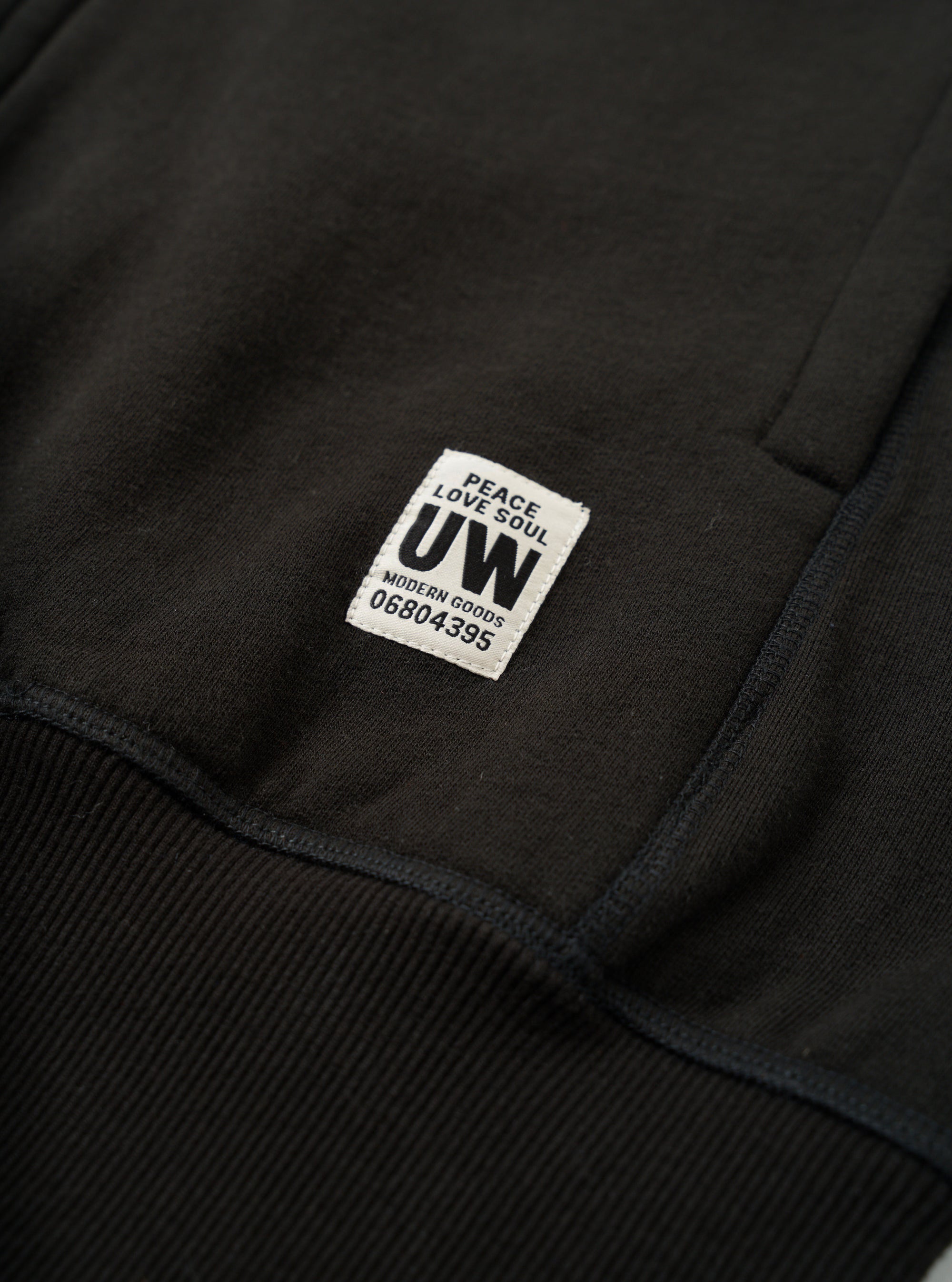 Close-up detail of a black brushback sweat core hoody, showing a woven brand label reading "Peace Love Soul UW Modern Goods 06804395" stitched onto the chest. Ribbed cuff trim is visible at the lower left. The jersey fabric texture is clearly visible against a dark studio backdrop.
