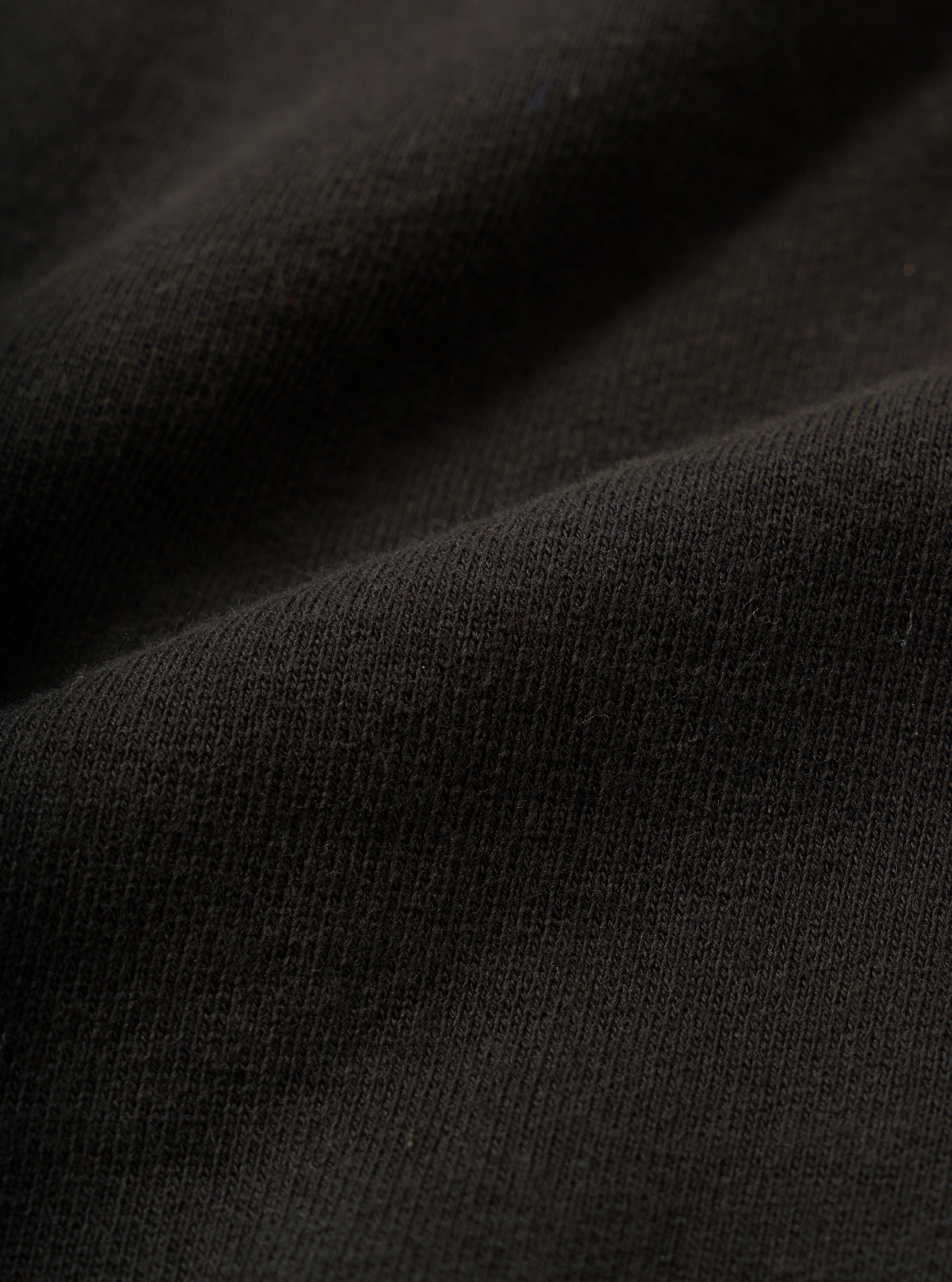 Close-up of black brushed-back sweatshirt fabric showing a fine knit jersey face with a soft, dense texture. The material is draped in loose folds, revealing its weight and handle. Shot against a dark background.