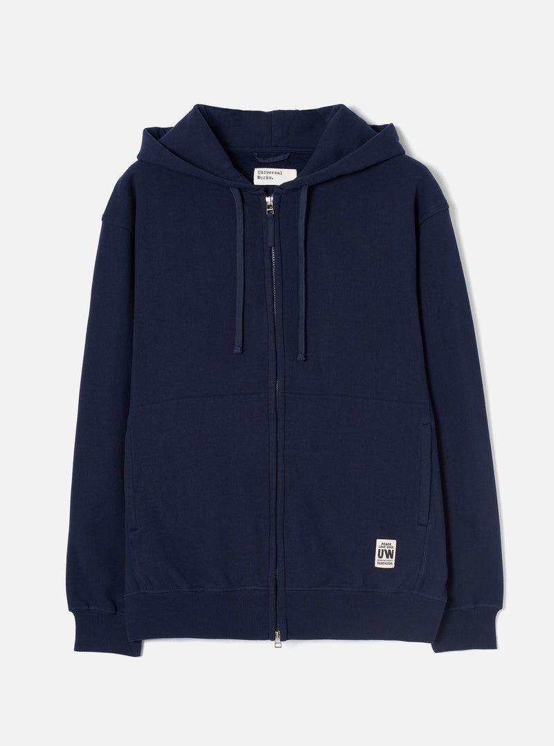 Navy brushback fleece zip-through hoodie laid flat on a white studio backdrop. Full-length silver metal zip, hood with fabric drawcords, two lower patch pockets, ribbed cuffs and hem. Small woven UW brand label at lower right. Universal Works neck label visible inside the hood.