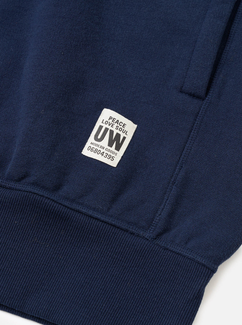 Close-up flat lay of a navy brushback hoodie, focusing on the lower front panel and ribbed hem. A woven label reading "Peace Love Soul UW Modern Goods 06804395" is stitched onto the body above the hem. A kangaroo pocket seam is visible to the upper right. The fabric is smooth jersey with a ribbed waistband below.