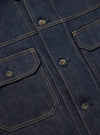 Close-up of the Indigo 13oz Selvedge Denim Trucker Jacket. Shows the indigo denim, silver buttons, and tan stitching on a flat, dark surface.