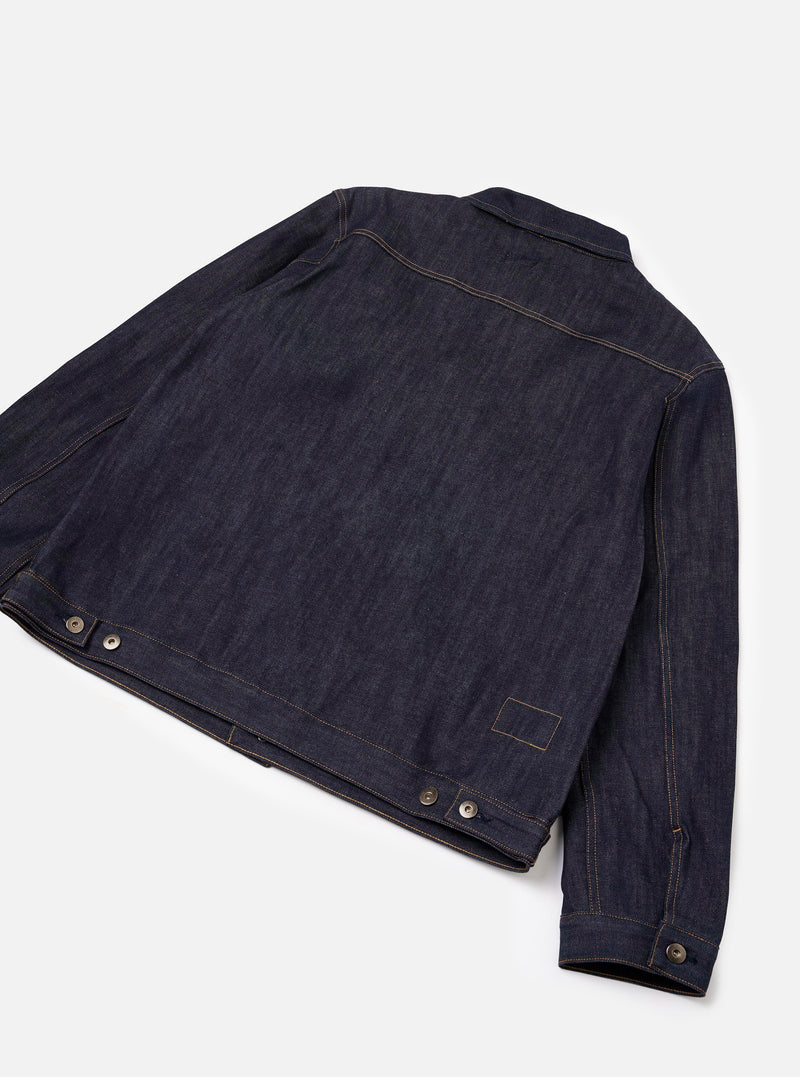 Rear perspective of the Indigo 13oz Selvedge Denim Trucker Jacket set against a neutral backdrop. Emphasizes the rich selvedge denim weave, panel seams across the shoulders and back, along with hem adjustment tabs at the sides.