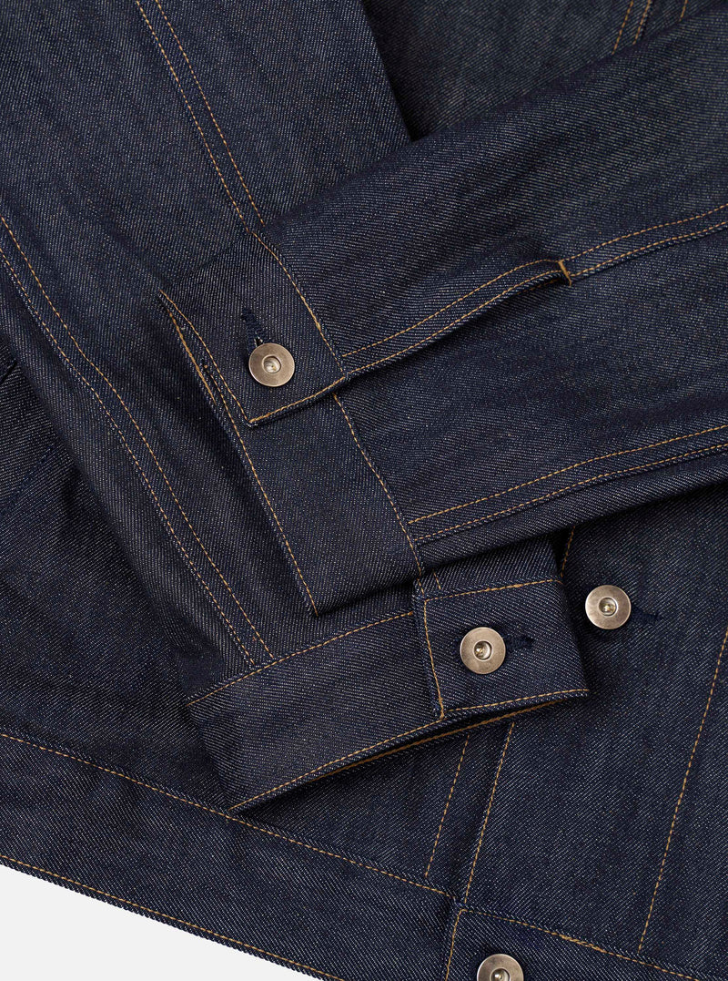 A detailed close-up shot of the Indigo 13oz Selvedge Denim Trucker Jacket on a plain studio backdrop. Focuses on the selvedge denim texture, silver metallic buttons, neat collar construction, and precise contrast seam stitching.