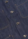 Close-up of the Indigo 13oz Selvedge Denim Bakers Jacket. Features include indigo denim, silver buttons, and tan stitching. The jacket is laid flat on a neutral surface.