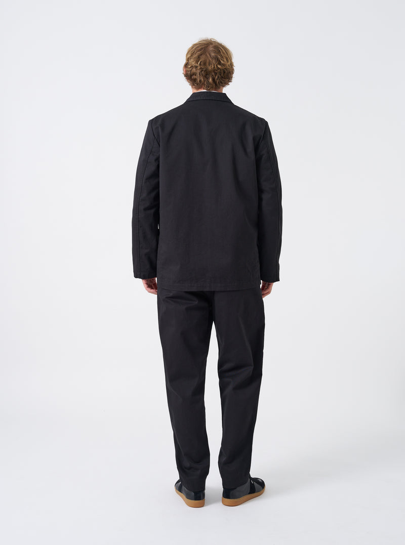 A male model wearing the Black Twill Three Button Jacket in an unbuttoned pose against a plain studio background, demonstrating how the jacket drapes over trousers and a layered shirt.