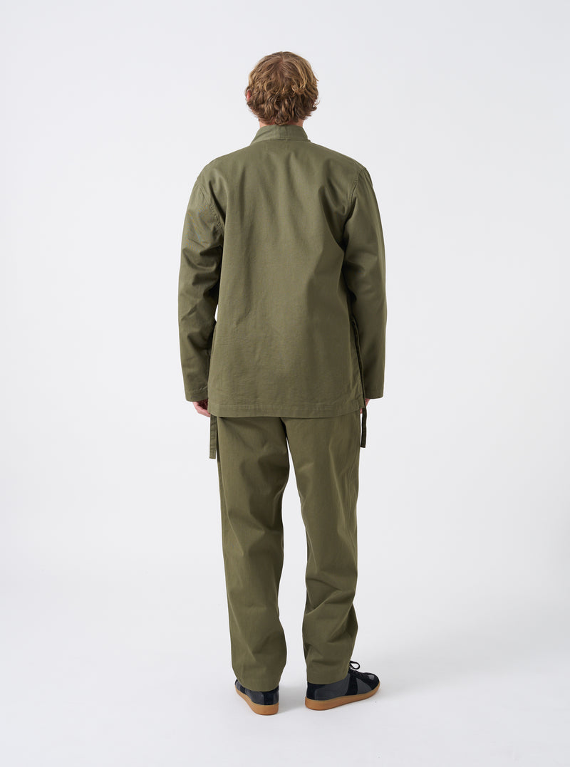 A male model is wearing the Light Olive Twill Kyoto Work Jacket in a front view studio shot against a white backdrop. He wears the jacket tied at the front over a casual top and trousers.
