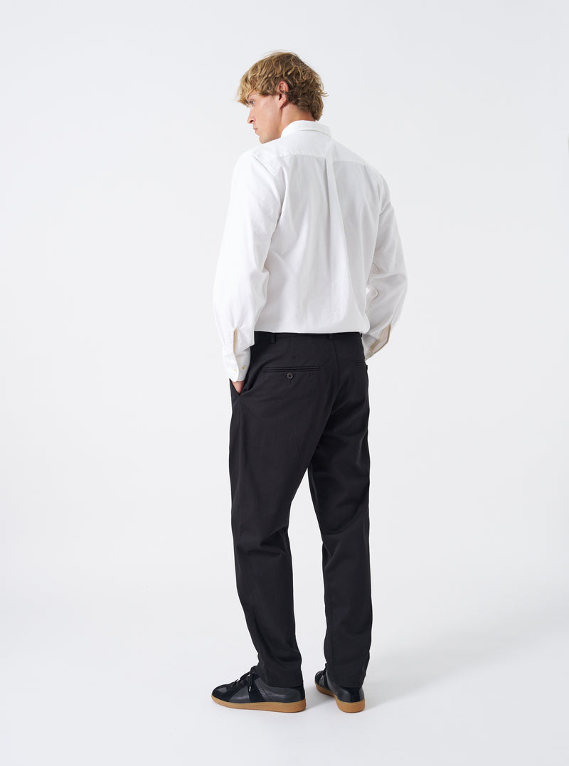 Rear view of a model wearing the Black Twill Military Chino against a plain white studio backdrop. Highlights how the dark cotton twill trousers fit through the seat and thighs, featuring rear jetted button pockets and clean hem finish.