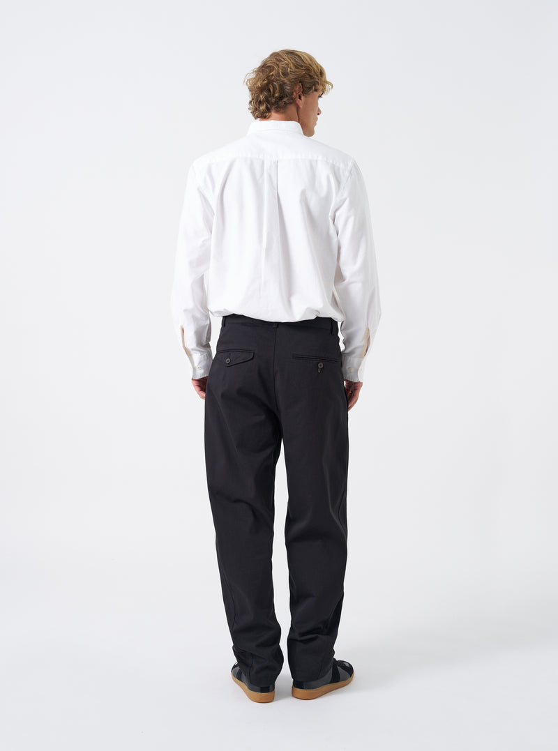 Rear view shot of a model wearing the Black Twill Curved Pant on a bright neutral backdrop. The model stands wearing the black trousers, highlighting the back pockets, waist fitting, and tapered leg opening at the ankles.