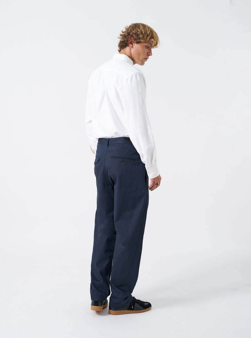 Rear view of a model wearing the Navy Twill Curved Pant against a plain white studio backdrop. The model wears the navy trousers, emphasizing the rear jetted and flap pockets alongside the signature curved silhouette.