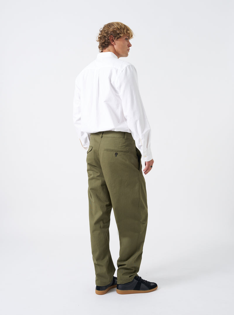 Back view of a model wearing the Light Olive Twill Curved Pant against a plain off-white studio backdrop. The model stands showing the rear button flap pocket detail, natural waist position, and unique silhouette.
