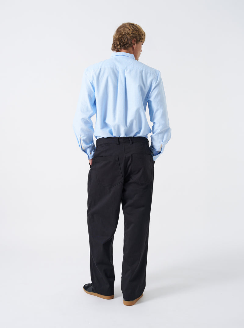 Back view shot of a model wearing the Navy Twill Duke Pant against a clean neutral studio backdrop. The model stands wearing the dark navy pants, demonstrating the relaxed drape, back patch pockets, and slight leg taper.
