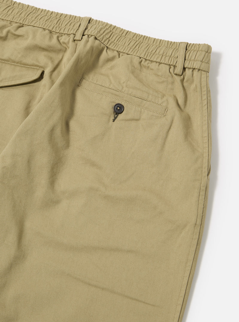 Close-up of the sand twill pleated track pant on a white surface. The pants are a light tan color and have a buttoned back pocket and elastic waistband.