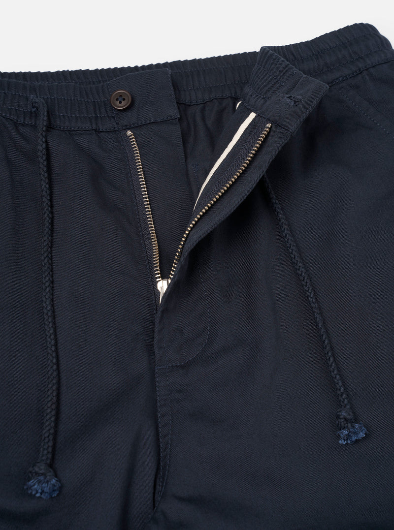 Close-up view of the Navy Twill Hi Water Trouser against a neutral studio backdrop. Highlights the texture of the 3/1 cotton twill weave, elasticated waistband, front drawstring, precise stitching, and slanted side hand pocket.