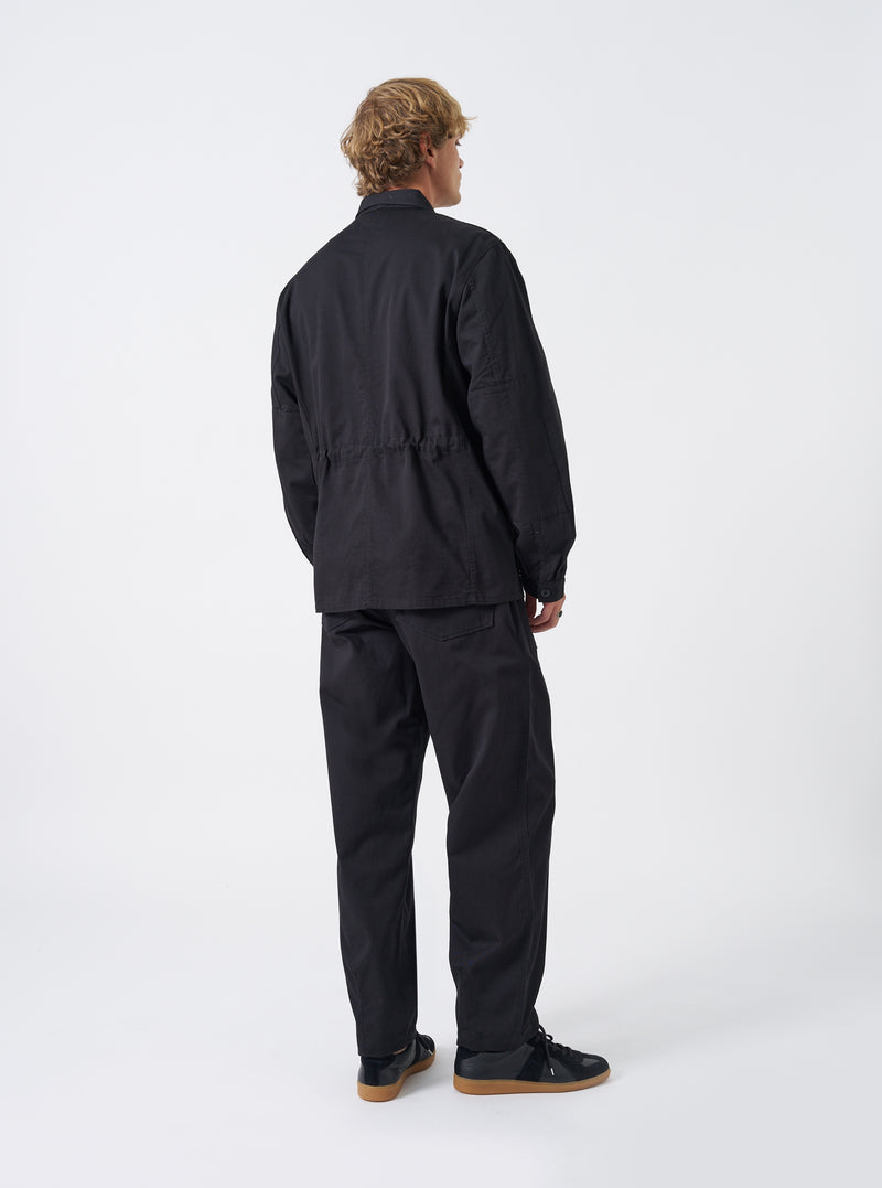 Rear view of a model wearing the Black Twill MW Fatigue Jacket against a plain neutral background. The photograph highlights the straight hemline, soft shoulder drape, structured twill fabric, and overall fit from behind.