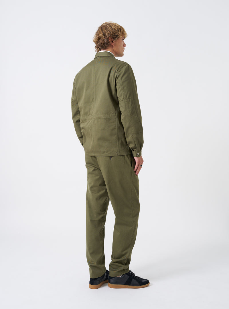 A model wears the Light Olive Twill MW Fatigue Jacket turned to show the side and back profile against a plain studio backdrop. The shot highlights the comfortable shoulder fit, sleeve cuffs, and hip-length hemline.