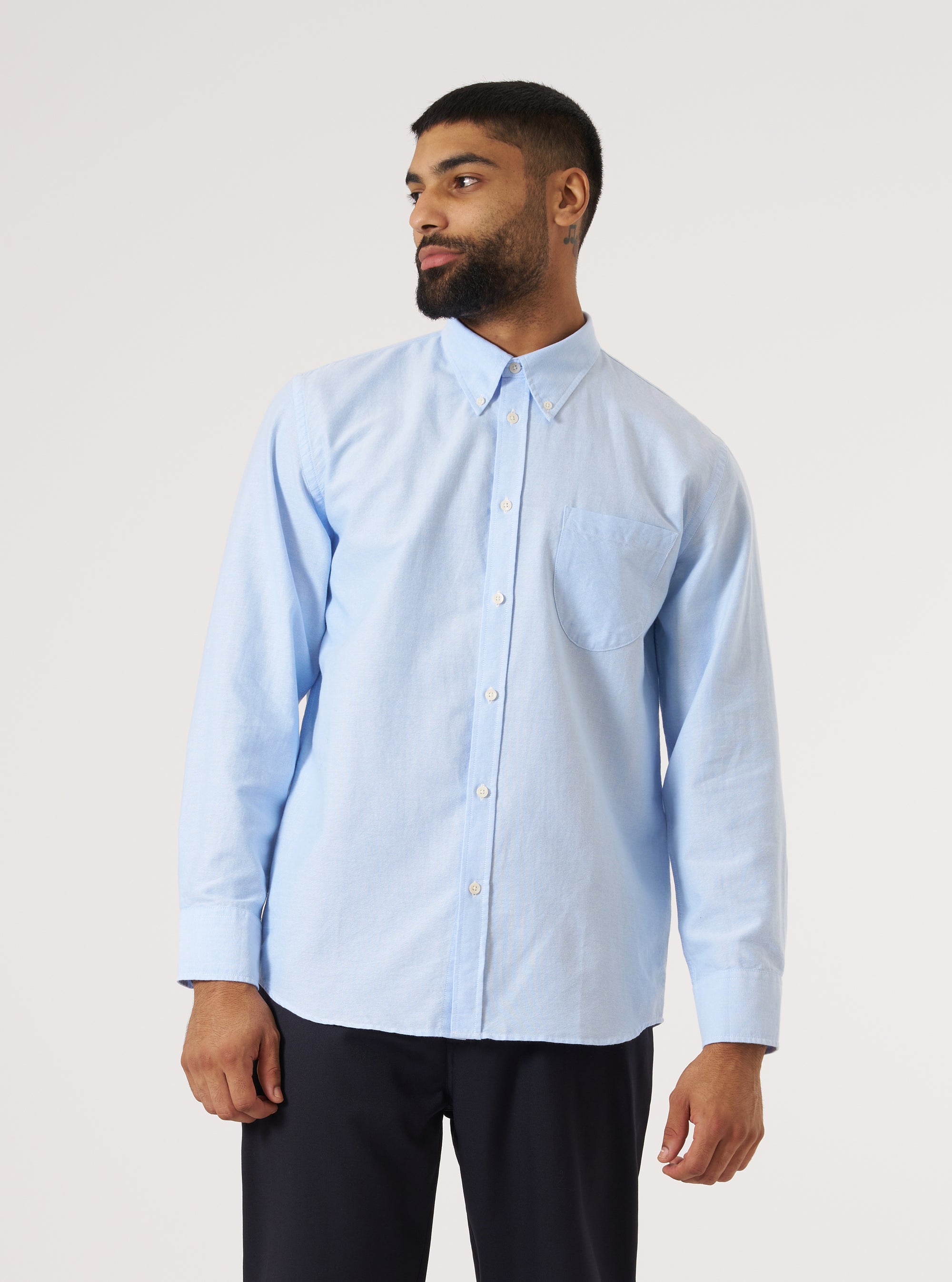 A man models the Sky Blue Oxford Daybrook Shirt in a studio setting. The light blue shirt is buttoned up and paired with dark pants against a white backdrop.