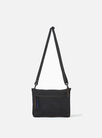 The master-piece x Universal Works Black Canvas Sacoche is shown as a flat lay against a plain white background. The small black shoulder bag features a main compartment with a zip closure, an adjustable shoulder strap with metal hardware, and a front slip pocket. A small collaborative logo patch is visible on the bottom corner.