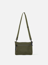The master-piece x Universal Works Olive Canvas Sacoche is shown as a flat lay against a white background. The small shoulder bag features a main zip compartment, a front zip pocket with two yellow zip-pullers, and a collaborative brand patch. It includes a thin, adjustable black crossbody strap attached with metal hardware.