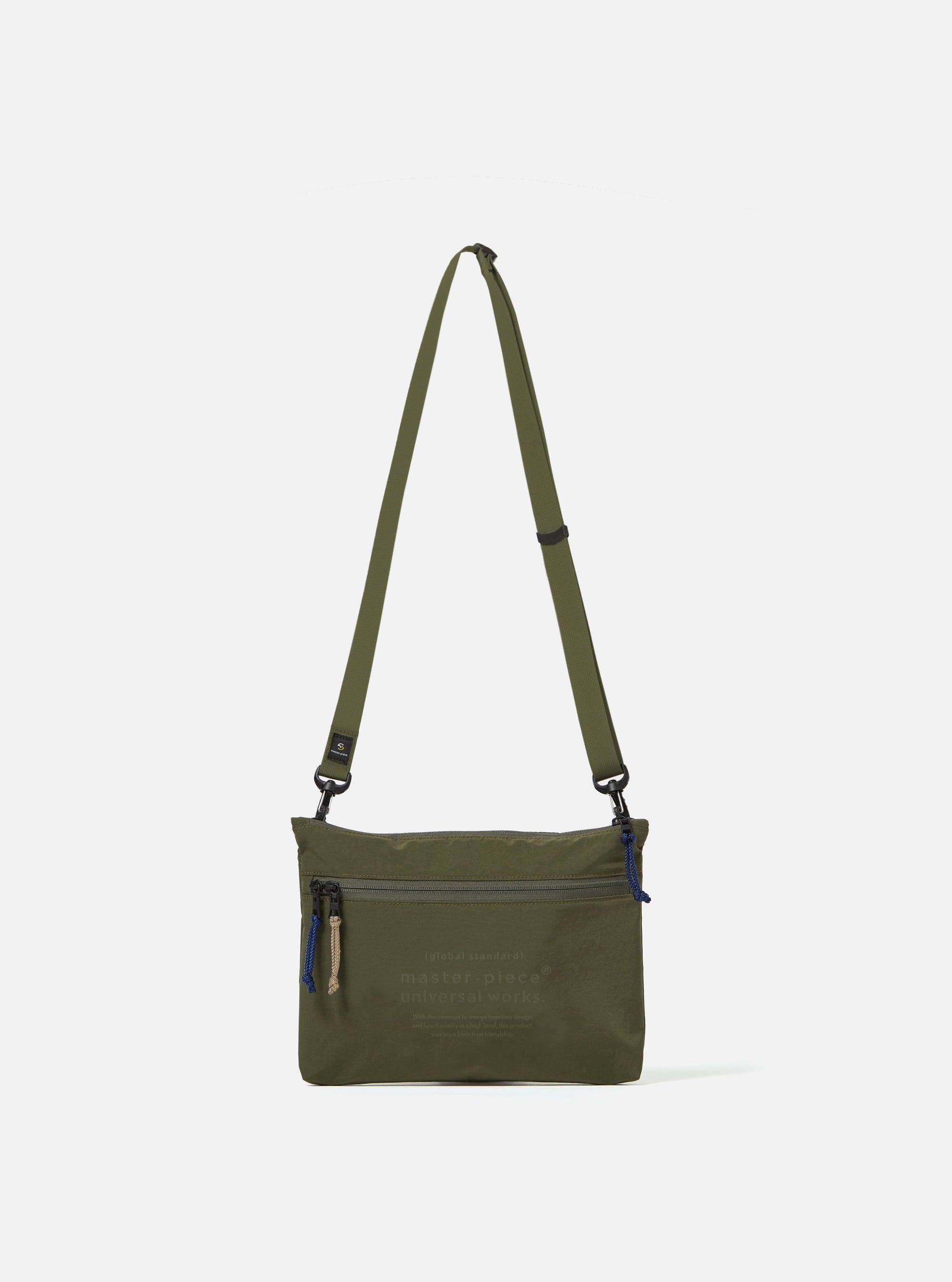 The master-piece x Universal Works Olive Canvas Sacoche is shown as a flat lay against a white background. The small shoulder bag features a main zip compartment, a front zip pocket with two yellow zip-pullers, and a collaborative brand patch. It includes a thin, adjustable black crossbody strap attached with metal hardware.