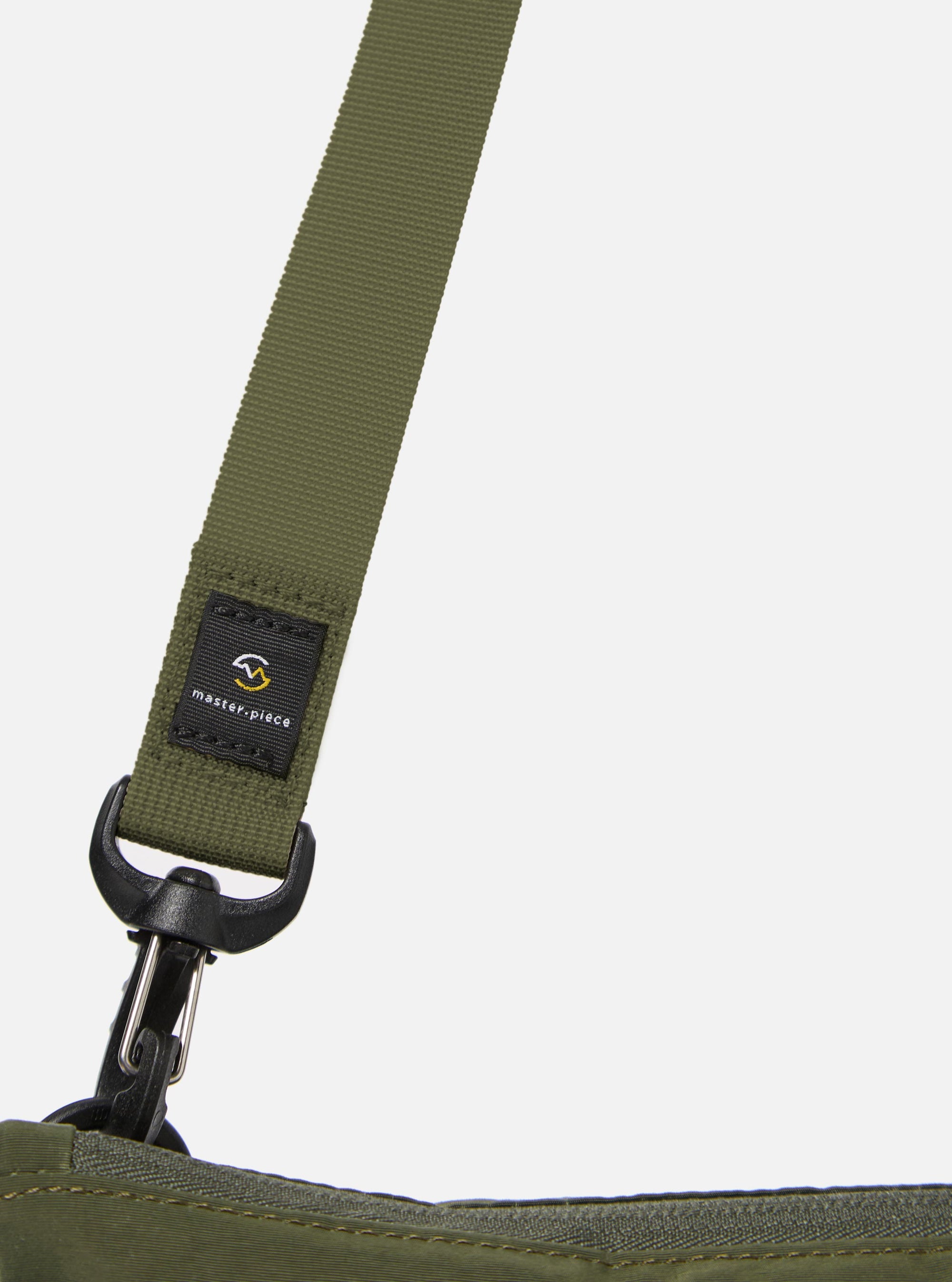 The master-piece x Universal Works Olive Canvas Sacoche is shown from the back as a flat lay against a plain white background. The image highlights the clean, rectangular construction of the olive green canvas bag and features a wide webbing strap at the rear, designed to attach to a suitcase handle.