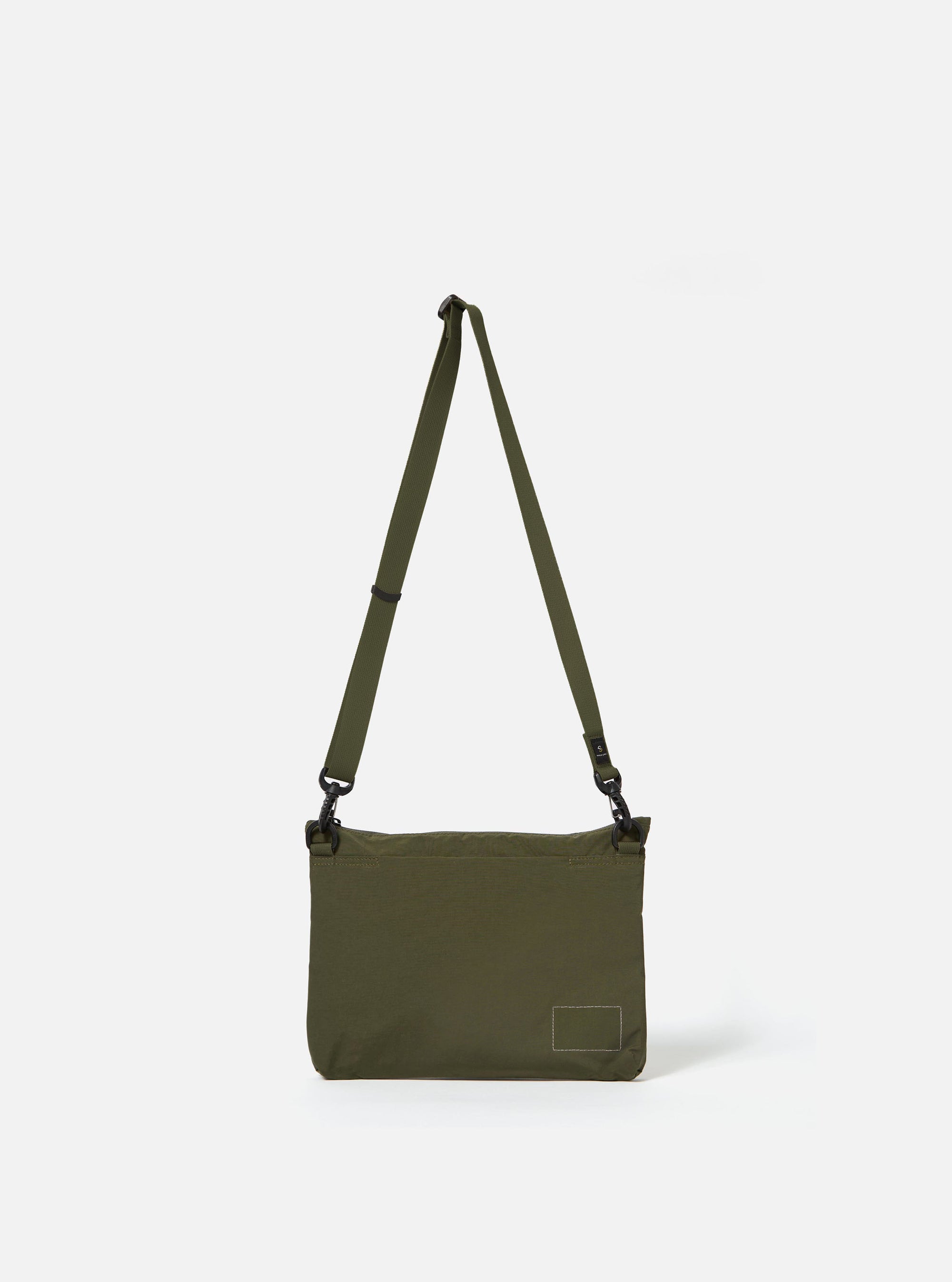 The master-piece x Universal Works Olive Canvas Sacoche is shown from a rear angle as a flat lay against a plain white background. The image highlights the back of the bag, featuring a small zippered pocket and the adjustable olive webbing strap. The durable olive green canvas fabric is the prominent feature.