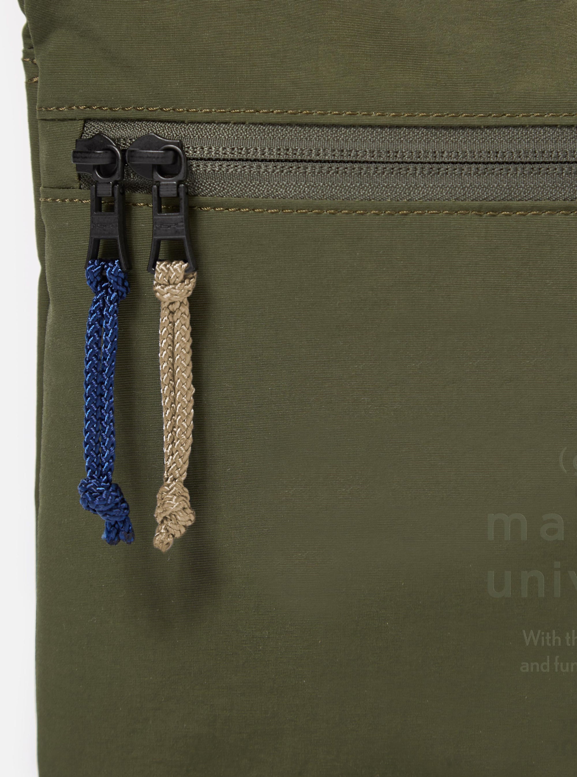 The master-piece x Universal Works Olive Canvas Sacoche is shown from a rear angle as a flat lay against a plain white background. The image highlights the back of the bag, featuring a clean olive green canvas surface and the attachment points for the thin, adjustable black crossbody strap.