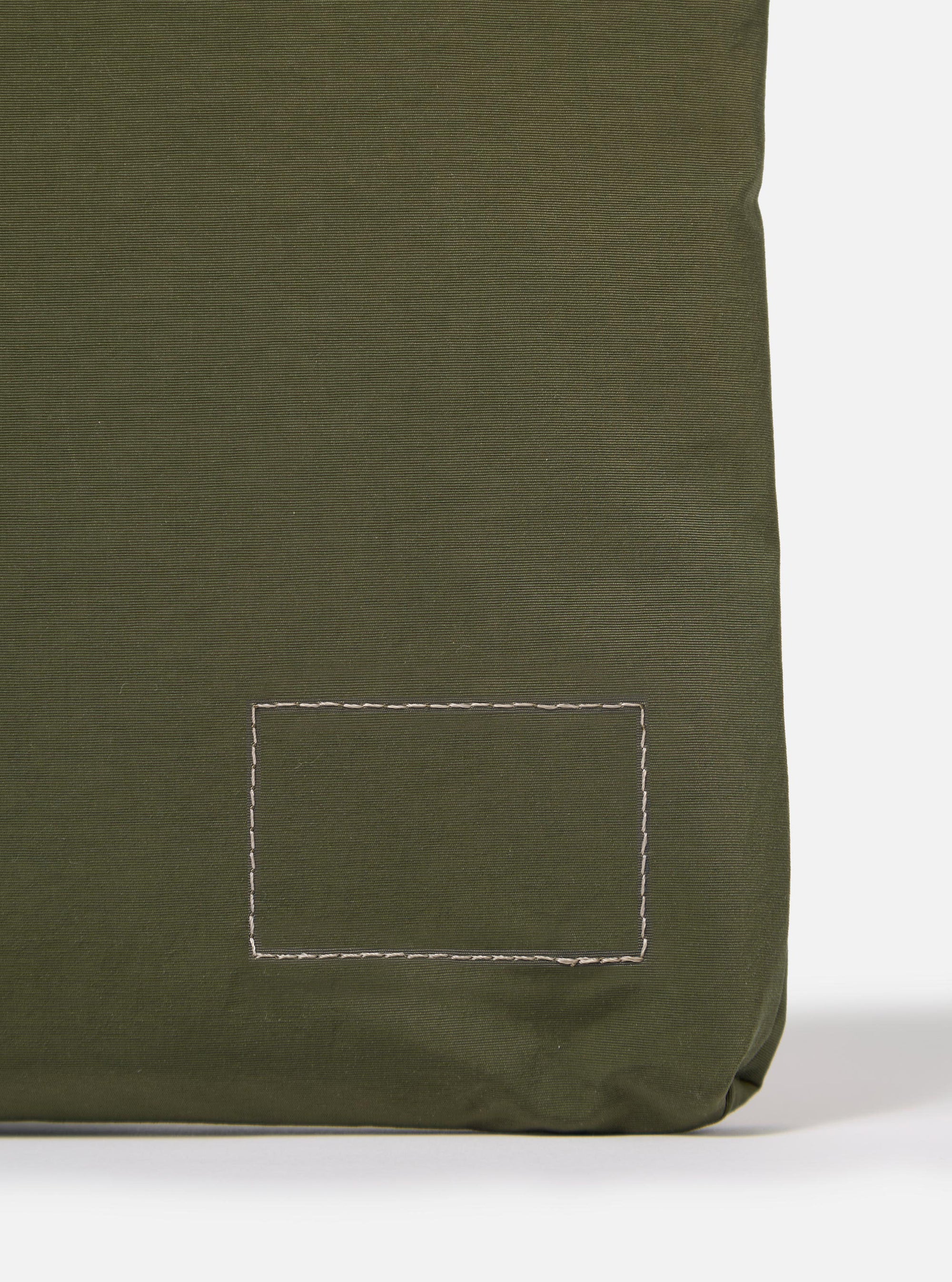 The master-piece x Universal Works Olive Canvas Sacoche is shown from a rear angle as a flat lay against a plain white background. The image highlights the back of the olive green bag, featuring a small woven brand label and the attachment points for the adjustable webbed shoulder strap.