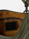 The master-piece x Universal Works Olive Canvas Sacoche is shown in a detail view as a flat lay against a plain white background. The image focuses on the side profile of the bag, highlighting the zippered main compartment, a side webbing loop with a branded master-piece tag, and the durable olive green canvas fabric.