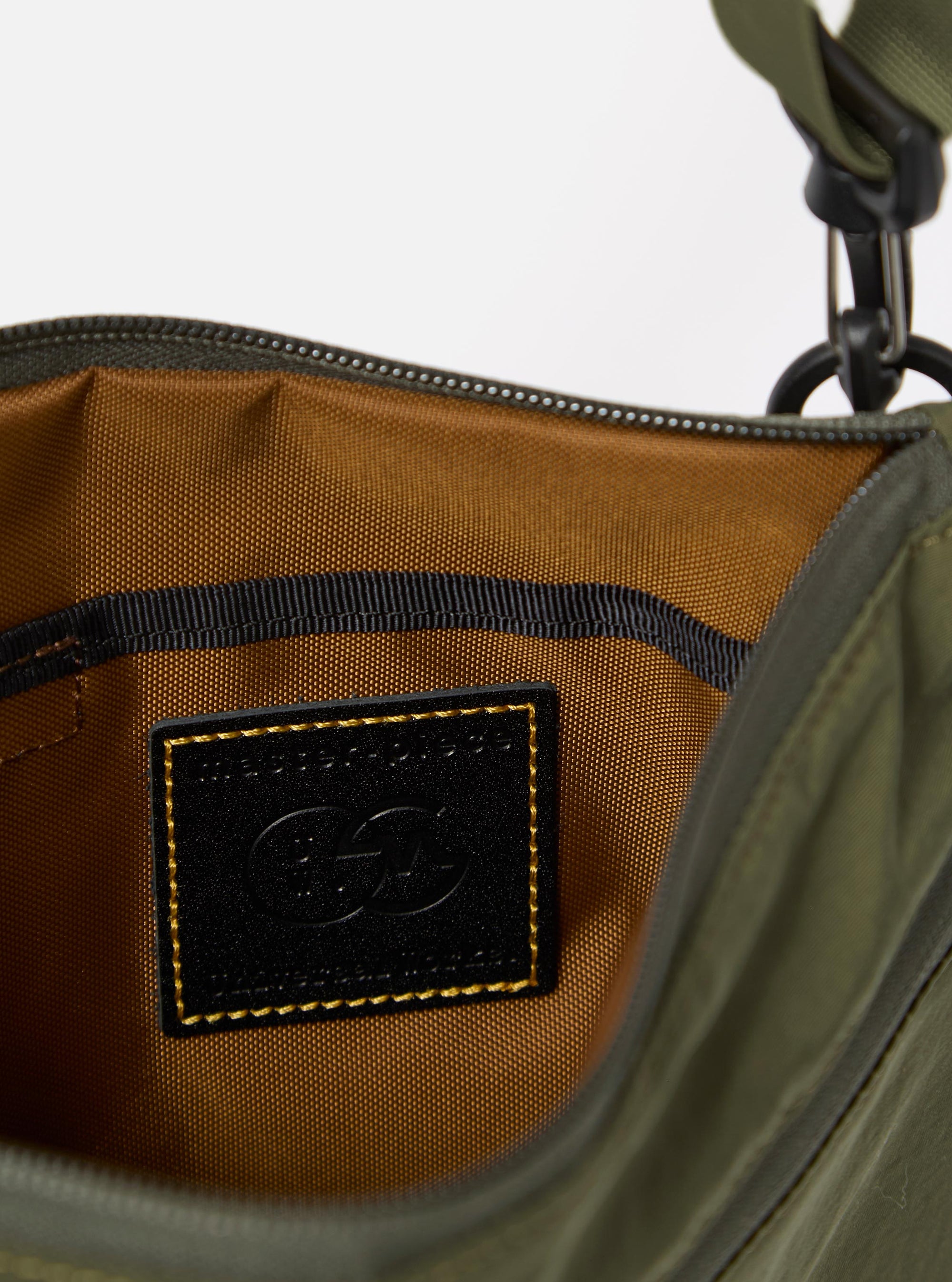 The master-piece x Universal Works Olive Canvas Sacoche is shown in a detail view as a flat lay against a plain white background. The image focuses on the side profile of the bag, highlighting the zippered main compartment, a side webbing loop with a branded master-piece tag, and the durable olive green canvas fabric.