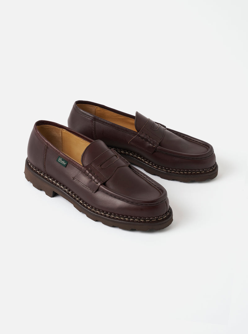 A pair of Paraboot Cafe Leather Reims Marche loafers rests on a light grey studio surface. Features a smooth dark brown leather upper with a penny strap across the vamp, tan lining, contrast braided welt stitching, and a chunky dark rubber lug sole.
