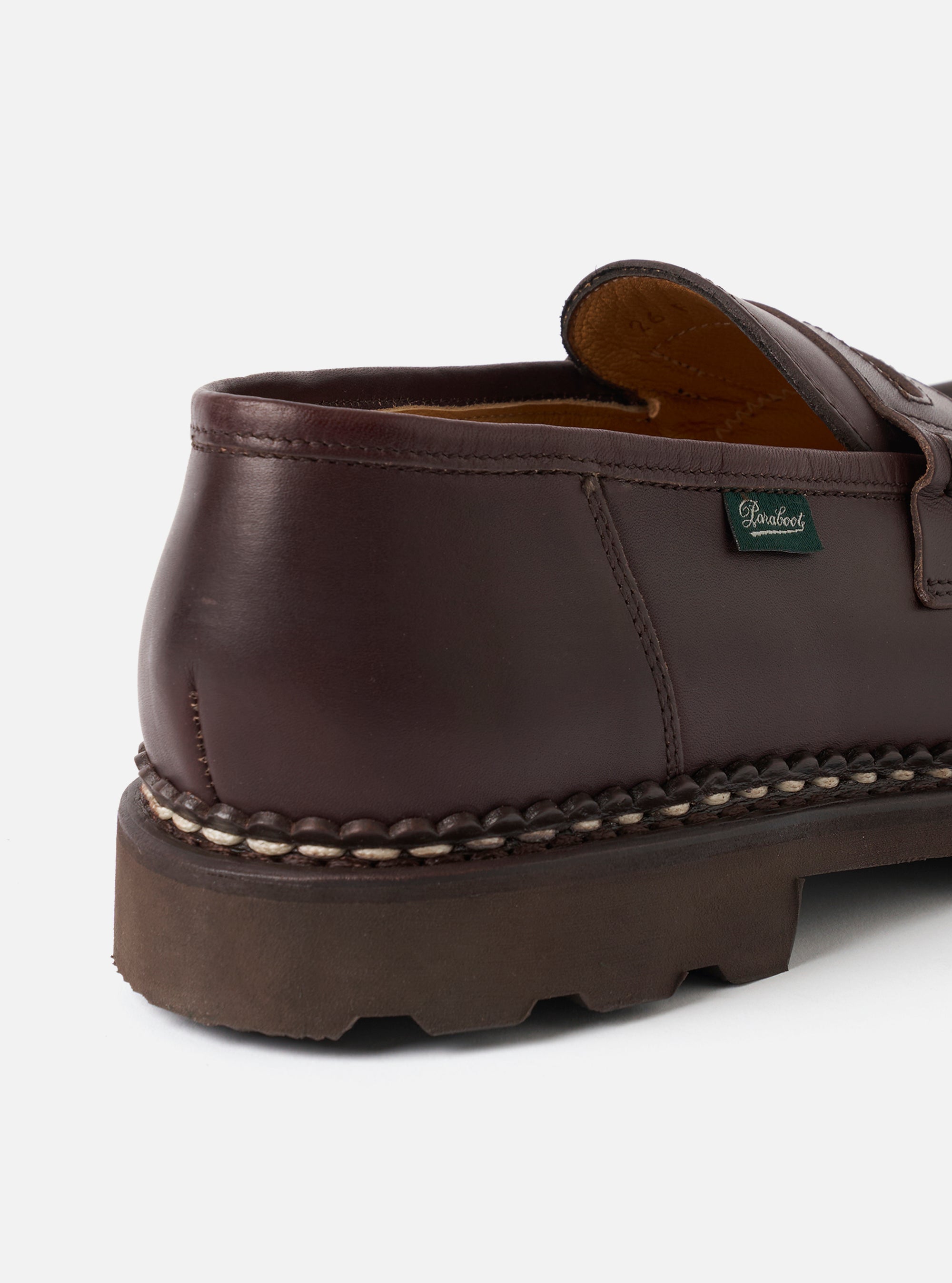 Close-up of the heel and counter of a dark brown leather loafer against a light grey studio backdrop. Visible details include a tan leather lining, a green Paraboot woven label on the heel counter, prominent cream Norwegian welt stitching, and a chunky dark brown rubber lug sole.