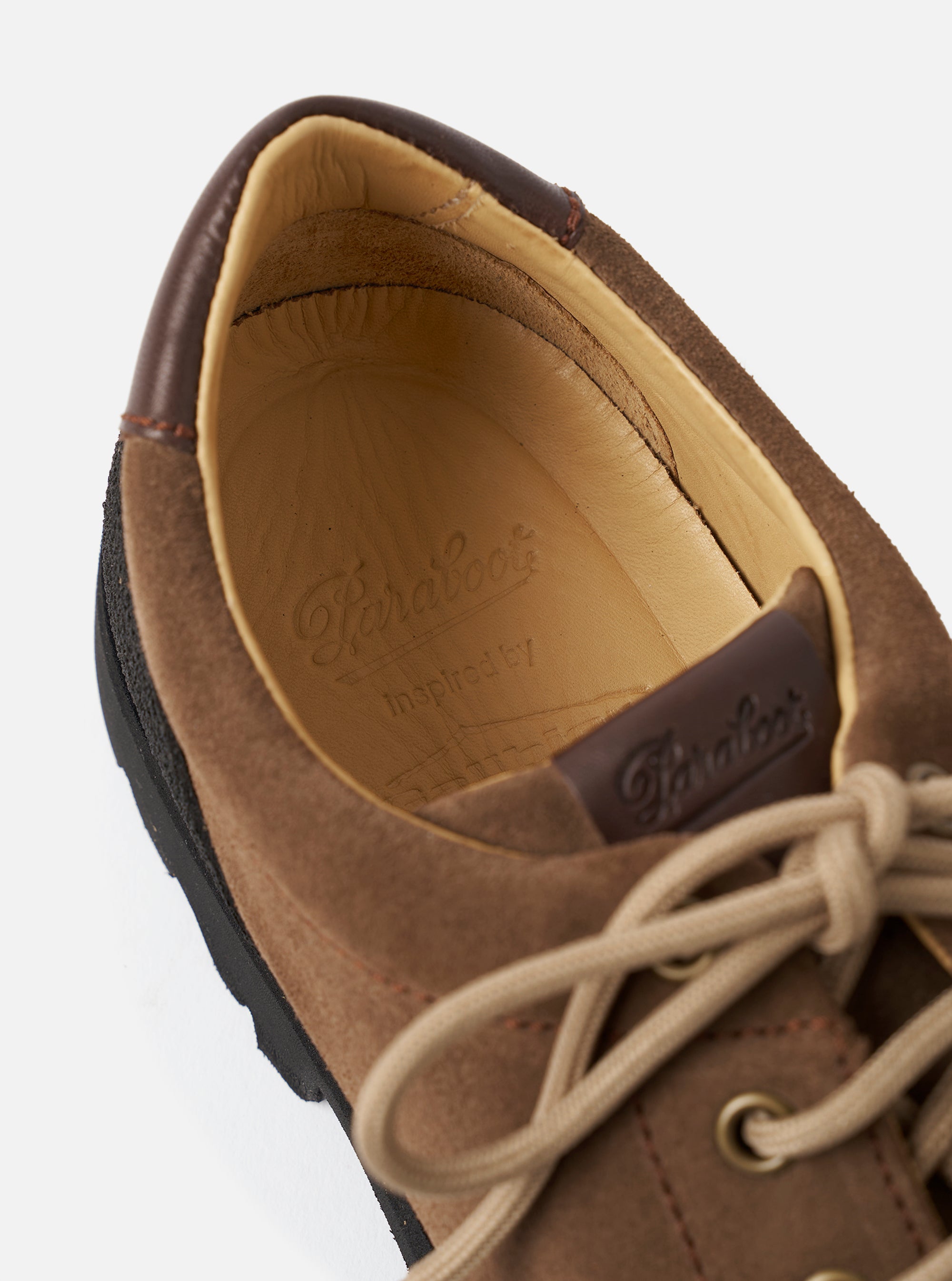 Close-up detail of the Paraboot Vel in marron suede, shot against a light grey studio backdrop. The image focuses on the open collar, revealing a tan leather insole stamped with the Paraboot logo and "Inspired by". A dark brown leather heel counter and branded pull tab are visible, with beige round laces threaded through metal eyelets. The chunky black rubber sole is visible at the edge.
