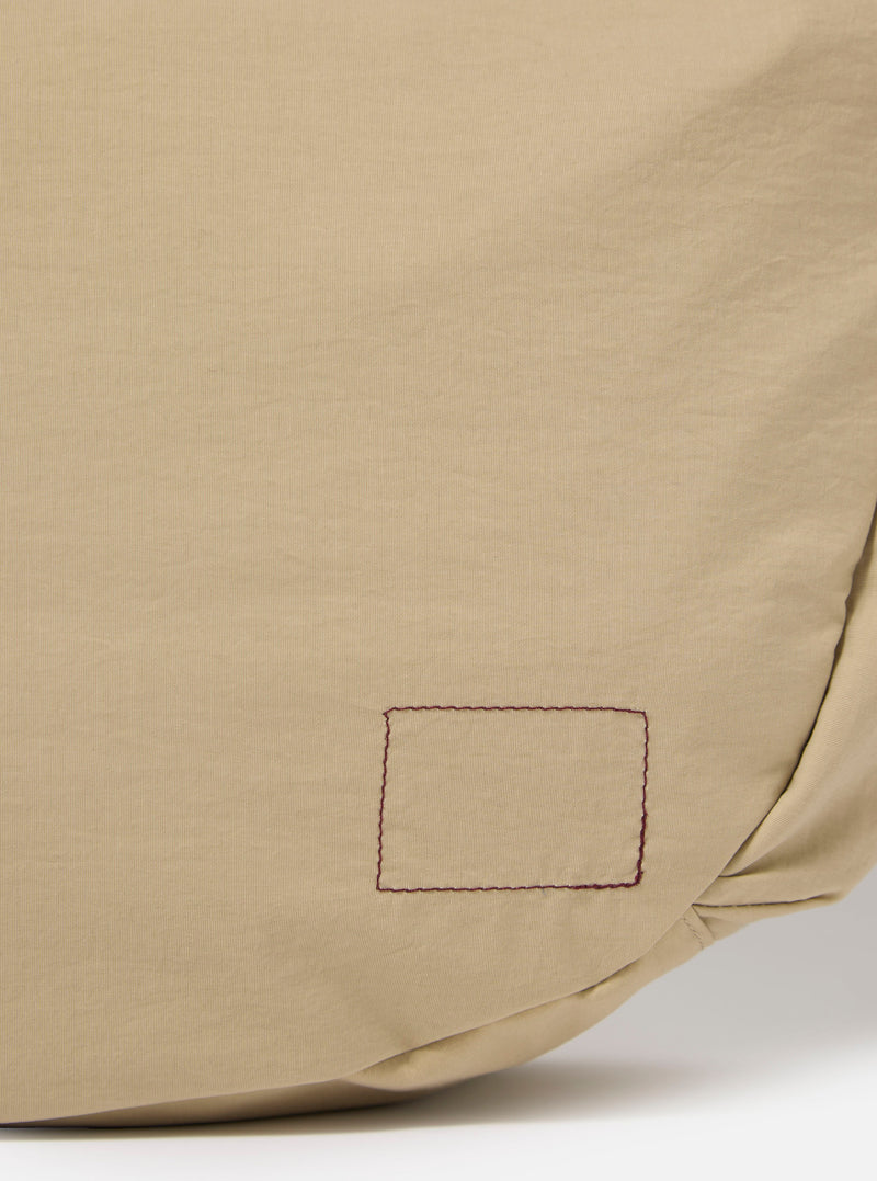 The master-piece x Universal Works Beige Large Canvas Shoulder Bag is shown from the back as a flat lay against a plain white background. The image highlights the adjustable webbed shoulder strap and the clean, rectangular construction of the bag's rear panel in a sand-colored beige fabric.