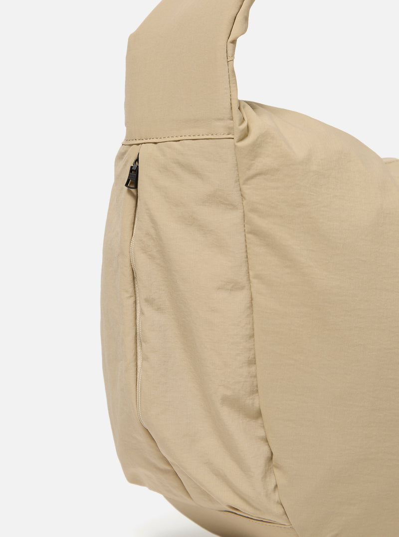 The master-piece x Universal Works Beige Large Canvas Shoulder Bag is shown from a side angle as a flat lay against a plain white background. The image highlights the depth of the bag, the sturdy canvas construction, and the adjustable shoulder strap. A small collaborative logo patch is visible on the side.