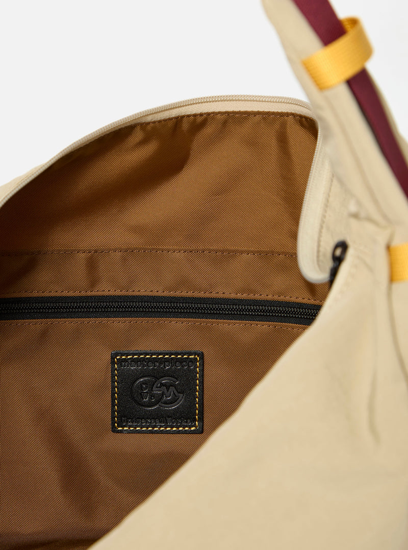 The master-piece x Universal Works Beige Large Canvas Shoulder Bag is shown from the back as a flat lay against a plain white background. The image highlights the wide, adjustable shoulder strap, a large external patch pocket with a dual-branded master-piece and Universal Works logo, and the durable beige canvas construction.