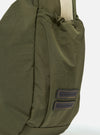 The master-piece x Universal Works Olive Large Canvas Shoulder Bag is shown from the back as a flat lay against a plain white background. The image highlights the rear of the bag, featuring a horizontal webbing strap designed for a suitcase handle and a tonal olive fabric. A small black brand tag is visible near the bottom.